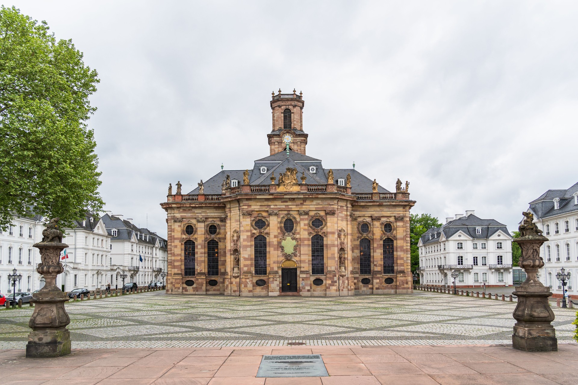 A large building Baroque church in the city of Saarbrucken in Germany with a clock tower and a small courtyard in front of it